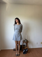 A woman stands in a room wearing a light gray long-sleeve mini dress with a flared skirt. She is posed in front of a plain beige wall with wood flooring, next to a small wooden stool holding a candle and a black-and-white patterned bag.
