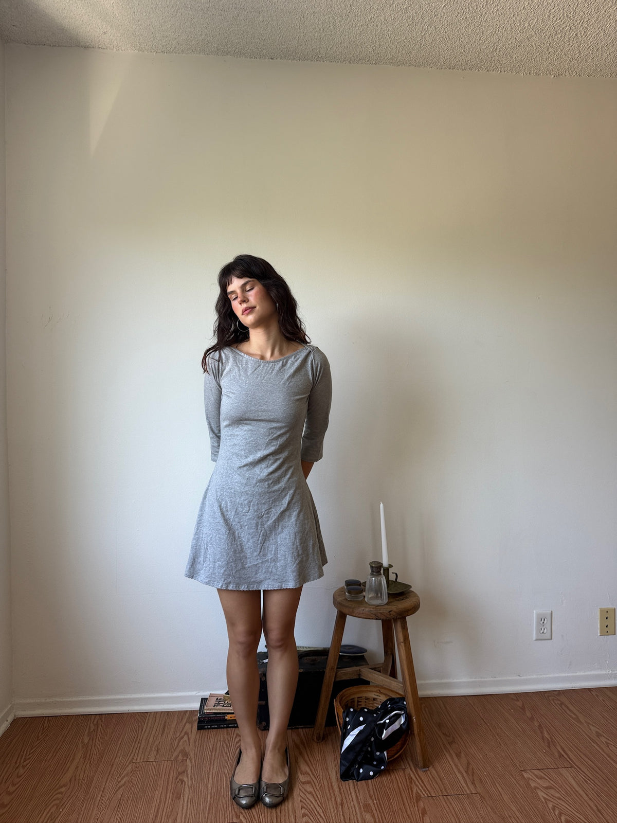 A woman stands in a room wearing a light gray long-sleeve mini dress with a flared skirt. She is posed in front of a plain beige wall with wood flooring, next to a small wooden stool holding a candle and a black-and-white patterned bag.