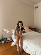 A woman sits on a bed in a bedroom, wearing a gray long-sleeve top, white shorts, and leopard print heels. The room has beige walls, a wooden floor, and white bedding.