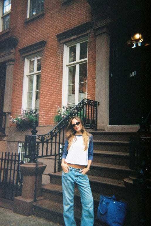 A woman wearing a white and navy raglan t-shirt, light blue wide-leg jeans, and sunglasses stands on the steps of a brick townhouse.
