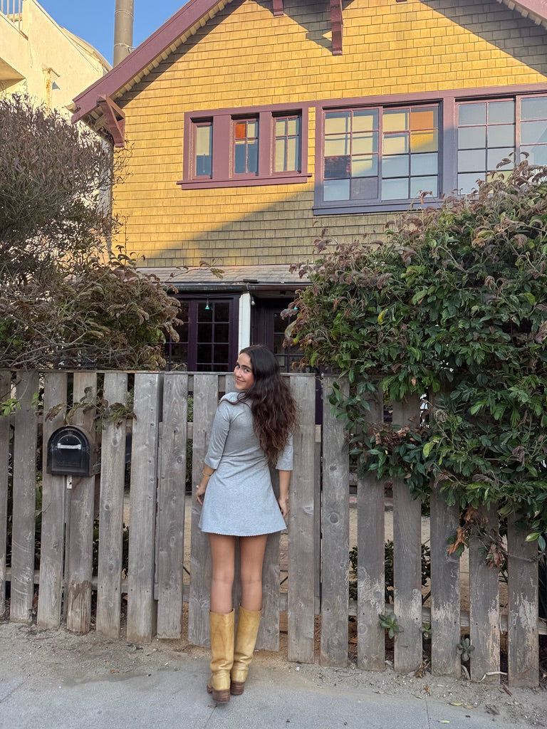 A woman stands outdoors in front of a wooden fence and yellow house, wearing a light blue mini dress and tan knee-high boots.