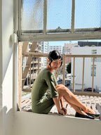 A woman wearing a short-sleeved olive green dress sits on a window ledge in a sunlit room, looking down and to the side. Outside the window, a city street scene with buildings and a railing is visible under a bright blue sky.