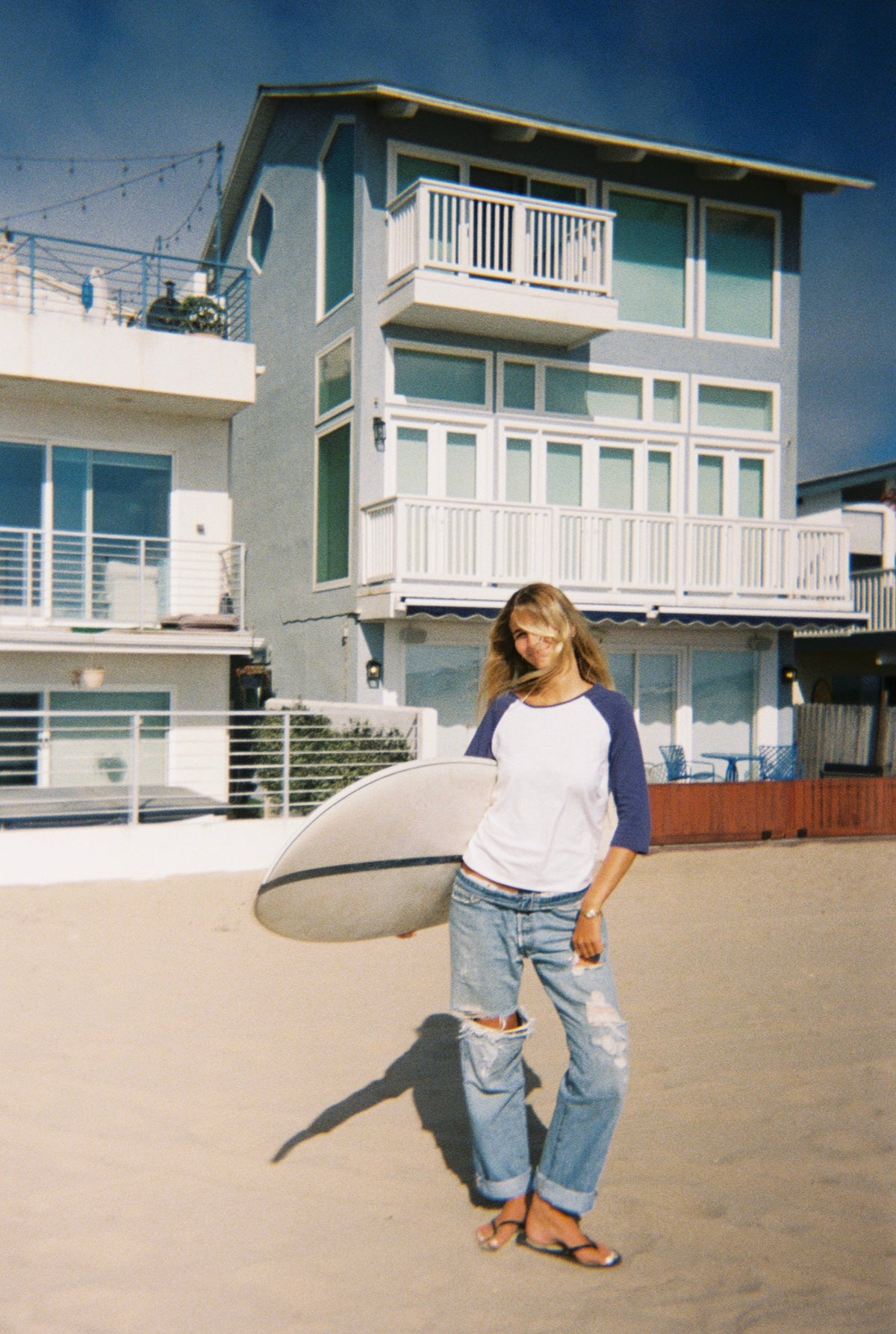 A woman stands on a sandy beach holding a surfboard, wearing a white and navy raglan t-shirt, ripped blue jeans, and flip-flops, with beachfront houses and a blue sky in the background.