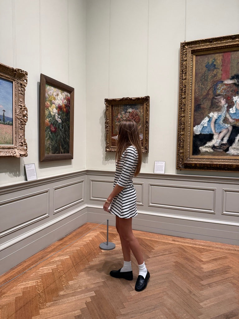 A woman in a black and white striped mini dress stands in a gallery room looking at framed paintings on the walls.