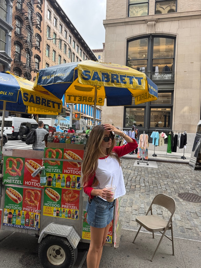 A woman wearing a white and red long-sleeve shirt, denim shorts, and sunglasses stands next to a street food cart with yellow and blue Sabrett umbrellas on a city street.