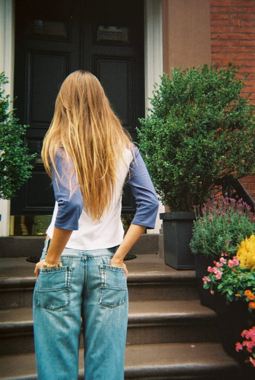 A woman with long blonde hair stands on stone steps in front of a building entrance, viewed from behind. She is wearing a white and blue raglan-style t-shirt and light blue jeans.