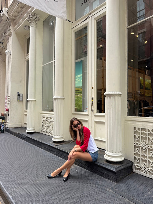 A woman wearing a red and white raglan t-shirt, denim shorts, and black flats sits on the black steps of a classic building with white columns.