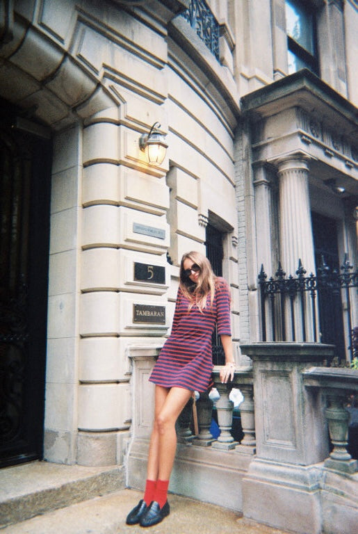 A woman posing on the steps of a grand stone building, wearing a short-sleeved purple and red striped mini dress, red socks, and black loafers.