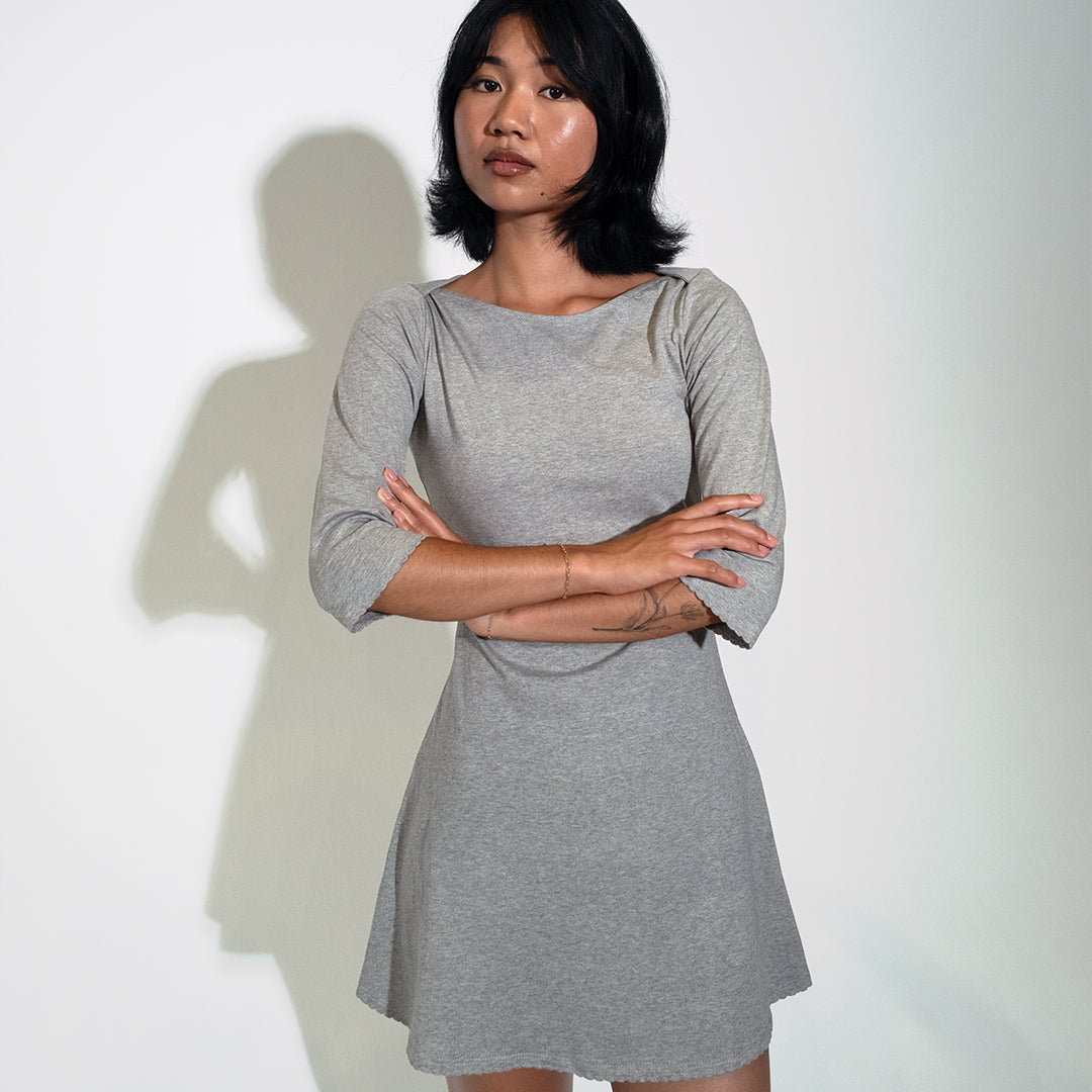 A woman wearing a heather gray three-quarter sleeve dress stands with arms crossed against a plain light background, casting a shadow.