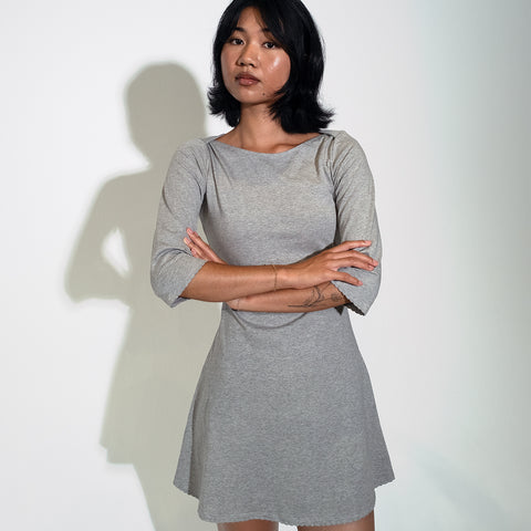 A woman wearing a heather gray three-quarter sleeve dress stands with arms crossed against a plain light background, casting a shadow.