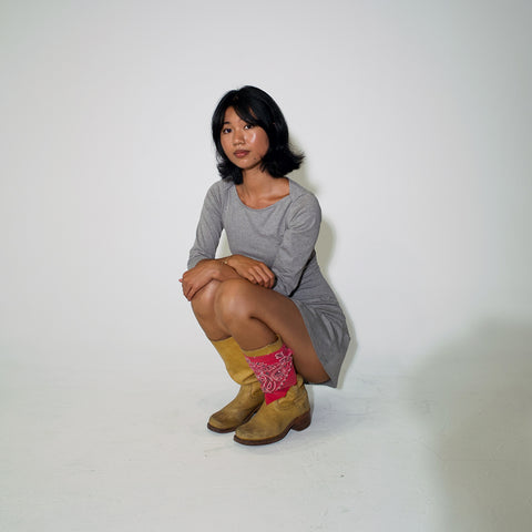 A woman crouching in a studio setting, wearing a gray long-sleeve dress, tan boots, and pink patterned socks against a plain white background.